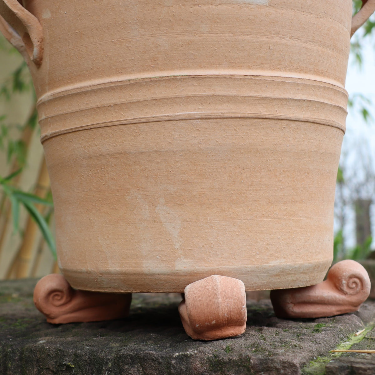 Ein Tontopf steht auf einer Steinoberfläche und wird von einem 4er-Set Topfschnecken aus Terrakotta erhöht, wobei jeder Block die Form einer gekräuselten Schnecke hat. Der Topf ist zylindrisch mit horizontalen Rillen und im Hintergrund ist teilweise Grün zu sehen.