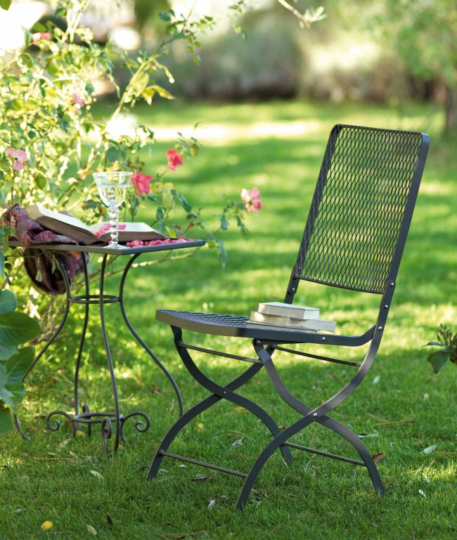 Ein Klappstuhl Aurora Gartenstuhl steht auf einem grünen Rasen neben einem kleinen Tisch. Auf dem Tisch stehen ein Glas Wasser, ein offenes Buch und eine Brille. Auf der Sitzfläche des Stuhls sind ein paar Bücher gestapelt. Im Hintergrund sind Büsche mit rosa Blüten zu sehen, die im Morgenlicht eine ruhige Atmosphäre schaffen.