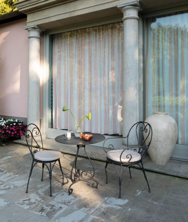 Ein kleiner schmiedeeiserner Tisch mit einer Kerze und einer Vase steht zwischen zwei passenden Armstühlen Aurora aus verzinktem Eisen mit weißen Kissen auf einer Steinterrasse vor Fenstern mit gestreiften Vorhängen. Neben dem Tisch steht ein großer Keramiktopf und in einem nahe gelegenen Gartenbeet blühen Blumen, die sich im Licht der Aurora sonnen.