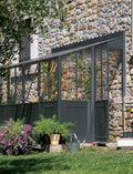 Eine Gartenszene mit einer Steinmauer im Hintergrund zeigt die Orangerie als Anbaumodell einer Hauswand mit transparenten Paneelen und einem dunklen Metallrahmen. Davor stehen Topfpflanzen verschiedener Größen, eine Gießkanne, eine Schubkarre und blühende Blumen. Im Vordergrund sind Gras und ein Busch zu sehen.