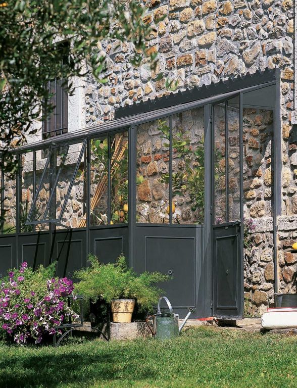 Eine Gartenszene mit einer Steinmauer im Hintergrund zeigt die Orangerie als Anbaumodell einer Hauswand mit transparenten Paneelen und einem dunklen Metallrahmen. Davor stehen Topfpflanzen verschiedener Größen, eine Gießkanne, eine Schubkarre und blühende Blumen. Im Vordergrund sind Gras und ein Busch zu sehen.