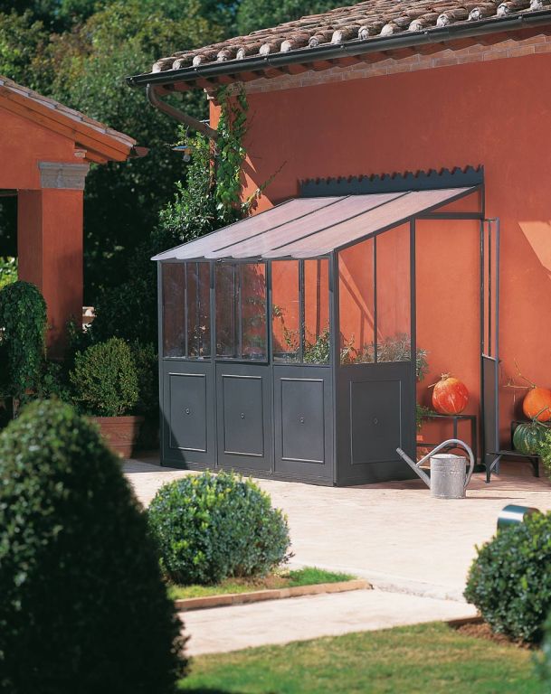 Die „Orangerie als Anbaumodell von einer Hauswand“, ein kleines Gewächshaus aus Metall und Glas, lehnt an einer rustikalen, orangefarbenen Stuckwand. Topfpflanzen umgeben den Bereich, davor steht eine Metallgießkanne, die ein Orangerie-Gefühl vermittelt. Die Terrasse ist sonnenbeschienen, und im Hintergrund ist Grün zu sehen, was auf eine üppige Gartenumgebung hindeutet.