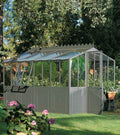 Ein üppiger, lebendiger Garten umgibt ein selbsttragendes Orangeriemodell mit grünem Rahmen und klaren Glasscheiben. In diesem Modell gedeihen verschiedene Pflanzen. Der Garten umfasst blühende Blumen, hohe Bäume und eine Atmosphäre der Ruhe und natürlichen Schönheit.