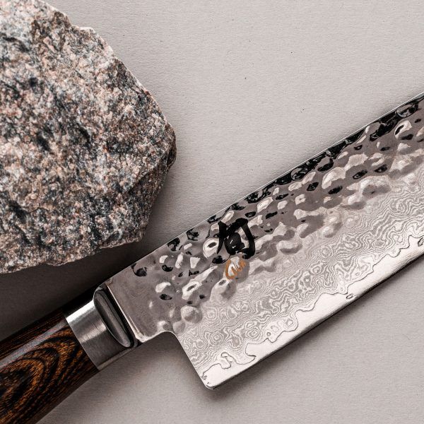 Eine Nahaufnahme des KAI Weihnachtsangebots 2-teiliges Geschenkset - Shun Premier Tim Mälzer Küchenmesser mit gehämmerter und gemusterter Klinge. Der Griff ist aus dunklem Holz. Das Messer liegt auf einer hellgrauen Oberfläche neben einem rau strukturierten Stein, perfekt für jedes Weihnachtsangebot oder Geschenkset, erhältlich in einer limitierten Auflage von 5.000 Sets.