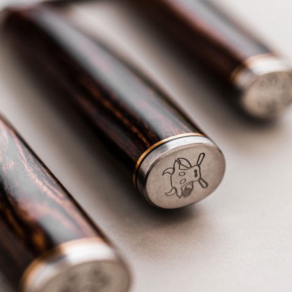 Nahaufnahme von drei Holzgriffen, jeder mit einer Metallendkappe, in die eine kleine Abbildung eines sitzenden Kaninchens eingraviert ist, das ein Blatt hält. Die Gravuren sind detailliert und die Holzmaserung ist sichtbar, was das edle Aussehen der Griffe unterstreicht – perfekt für jedes KAI Weihnachtsangebot 2-teiliges Geschenkset – Shun Premier Tim Mälzer – limitiertes 5.000 Stk. Set.