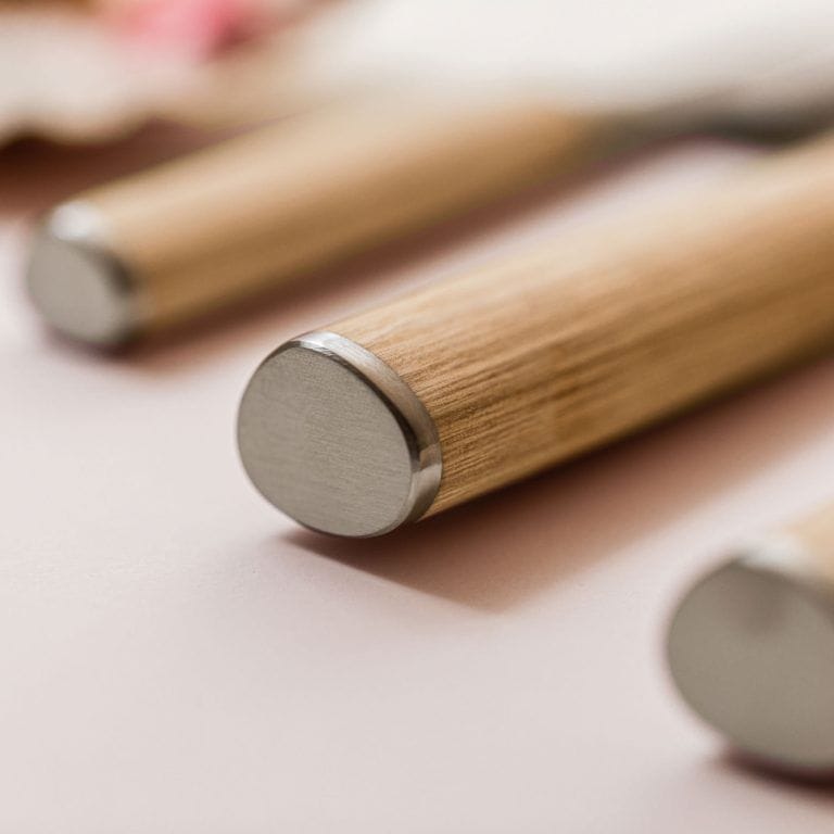 Nahaufnahme der Griffe von drei Küchenmessern, darunter das KAI X-MAS SELECTION DREAMBOX Shun Classic White Santoku, auf einer hellrosa Oberfläche. Die Griffe sind aus glattem Holz gefertigt und haben an den Enden Edelstahlkappen, die die Textur und das Material dieser exquisiten Stücke hervorheben.