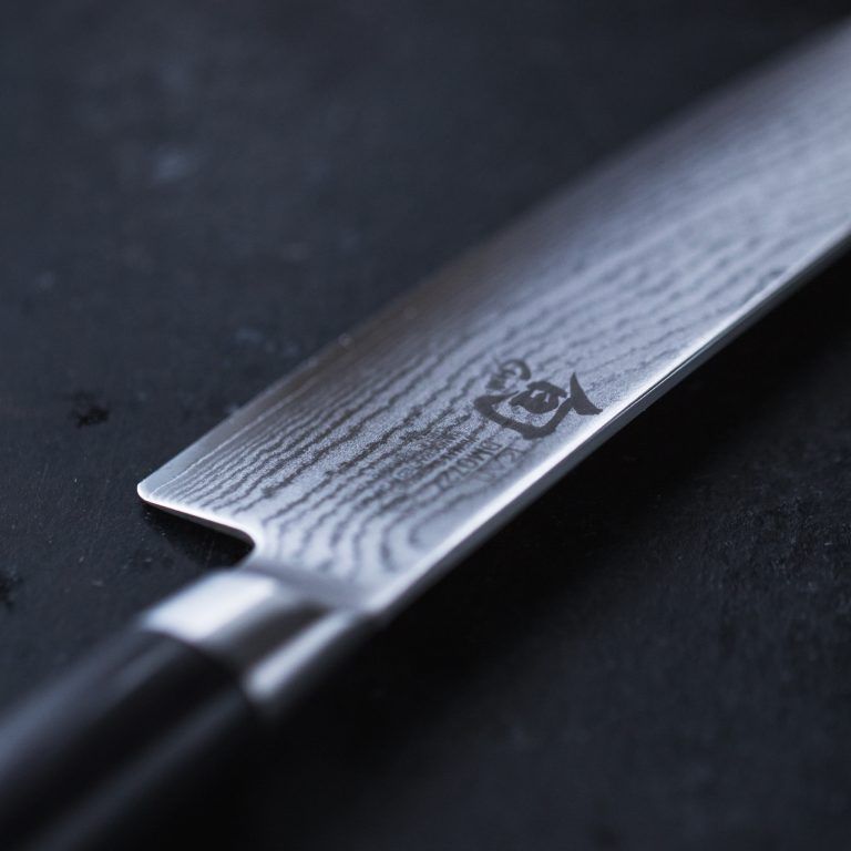 Nahaufnahme eines hochwertigen Küchenmessers aus dem 2-teiligen Geschenkset „KAI Weihnachtsangebot“ – Shun Classic-Serie, mit einer Klinge aus Damaszenerstahl mit komplizierten Mustern und einem dunklen Griff. Die fachmännische Handwerkskunst und die hochwertigen Materialien werden in der detaillierten Textur des Stahls deutlich, die sich vor einem dunklen Hintergrund abhebt.
