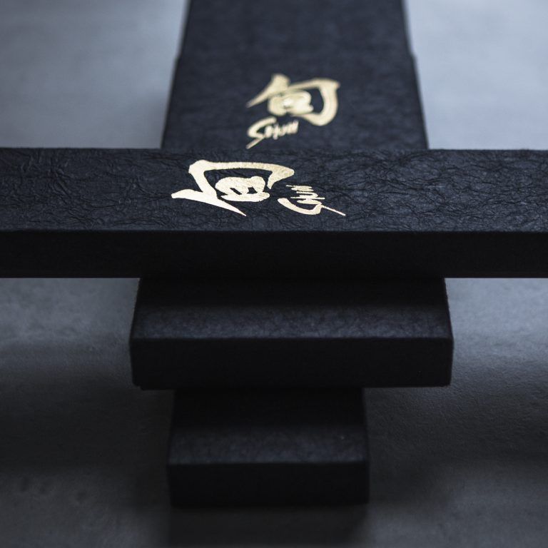 Nahaufnahme des 2-teiligen Geschenksets „KAI Weihnachtsangebot – Shun Classic“ mit schwarzem Design und markanten goldenen japanischen Schriftzeichen in der Mitte. Die strukturierte Oberfläche verstärkt das elegante Erscheinungsbild und macht es zu einer exquisiten Wahl für ein Weihnachtsangebot. Der Hintergrund ist unscharf, um sicherzustellen, dass die volle Aufmerksamkeit auf dieses Set in limitierter Auflage von nur 1.200 Stück gerichtet ist.