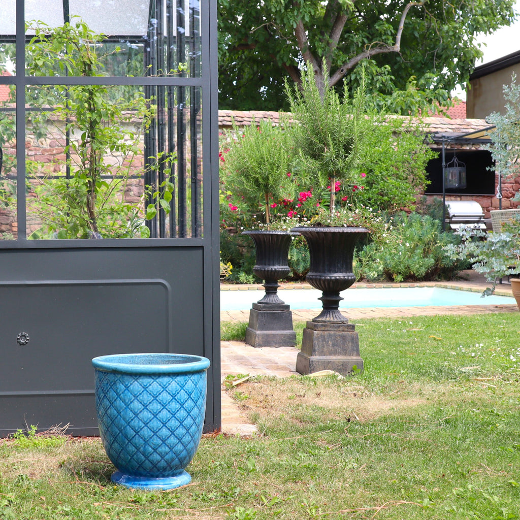 Ein Türkis Glasierter Blumentopf "Azur Provence" (Höhe 30,5-54 cm) steht auf dem Rasen nahe einem Gewächshaus mit Glaswänden. Im Hintergrund sind zwei schwarze Urnen, ein Pool, Bäume, Sträucher und eine Backsteinwand zu sehen.