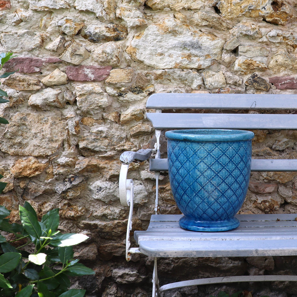 Der türkis verglaste Blumentopf "Azur Provence" (Höhe 30,5-54 cm) steht auf einer verwitterten grauen Metallbank vor einer rustikalen Steinmauer; unten links sind grüne Blätter und weiße Blumen zu sehen.
