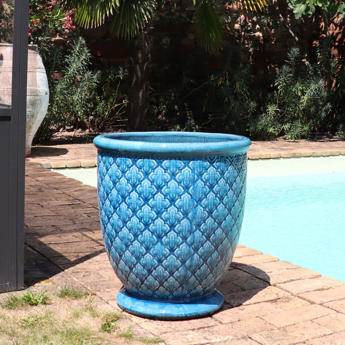 Der Türkis Glasierter Blumentopf "Azur Provence" (Höhe 30,5 bis 54 cm) mit Rautenstruktur steht auf einer gemauerten Terrasse am Pool, umgeben von üppigem Grün und eingefasst von einer Ziegelmauer.