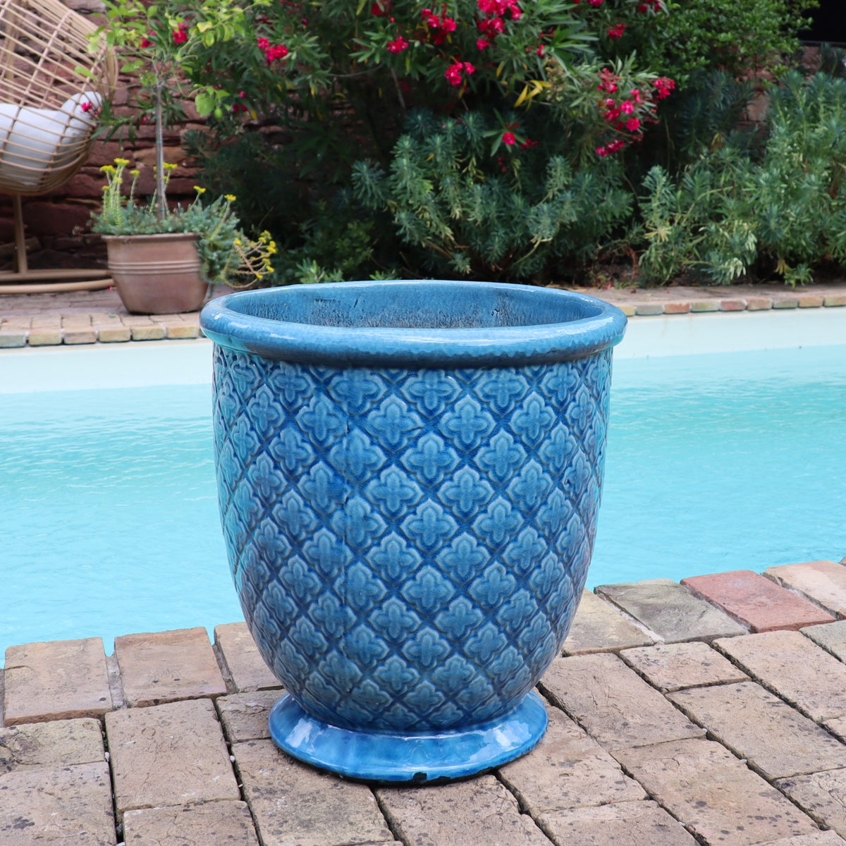 Der Türkis Glasierter Blumentopf "Azur Provence" (Höhe 30,5 bis 54 cm) mit einem strukturierten Muster steht auf einer gemauerten Terrasse am Pool, umgeben von üppigen grünen Pflanzen und leuchtend roten Blumen im Hintergrund.