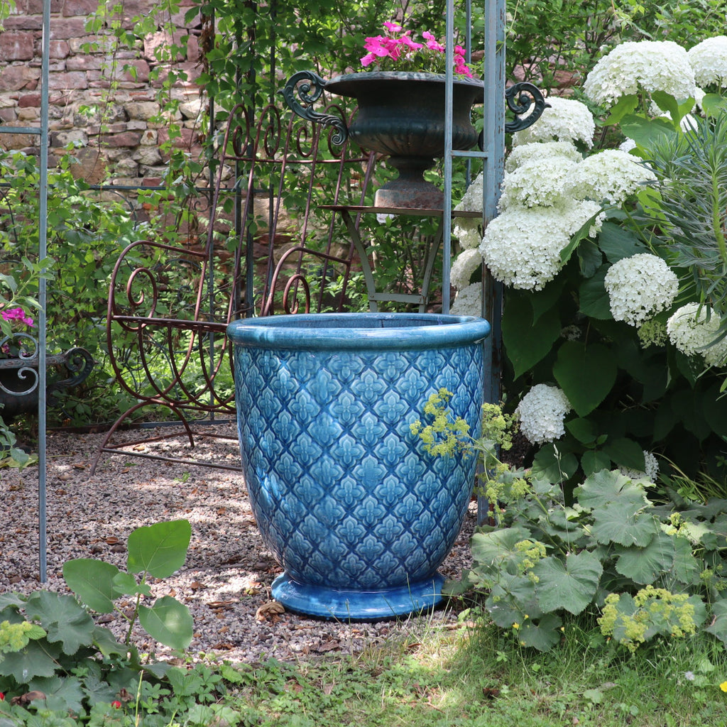 Der türkis glasierte Blumentopf "Azur Provence" (Höhe 30,5 bis 54 cm) steht auf Kies in einem üppigen Garten mit weißen Hortensien, grünem Laub, einem pinken Blumentopf und einer verzierten Metallbank im Hintergrund.
