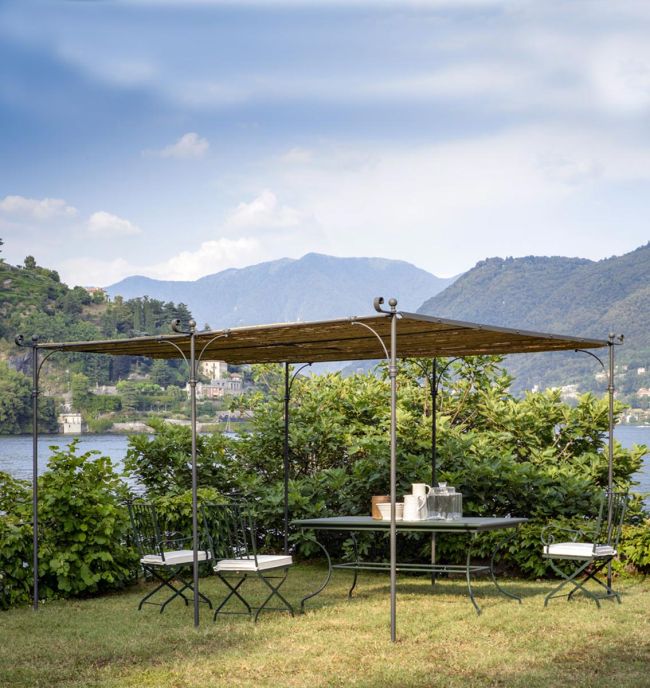 Eine malerische Außenanlage mit einer freistehenden linearen Pergola Solaire mit Metallrahmen und Baldachin, in der Nähe eines Sees gelegen. Unter der Pergola steht ein Tisch mit Stühlen und ein Tablett mit Getränken. Grüne Büsche umgeben den Bereich, im Hintergrund sind Berge und Gebäude zu sehen.