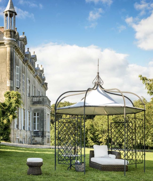 Eine malerische Szene eines großen Anwesens mit einem historischen Gebäude mit mehreren Schornsteinen und kunstvollen Details. Im Vordergrund steht ein stilvoller Pavillon Solaire rund mit weißem Baldachin und Korbmöbeln, darunter ein kleiner runder Korbtisch und ein Hocker, auf einem üppigen grünen Rasen.