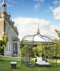 Eine malerische Szene eines großen Anwesens mit einem historischen Gebäude mit mehreren Schornsteinen und kunstvollen Details. Im Vordergrund steht ein stilvoller Pavillon Solaire rund mit weißem Baldachin und Korbmöbeln, darunter ein kleiner runder Korbtisch und ein Hocker, auf einem üppigen grünen Rasen.