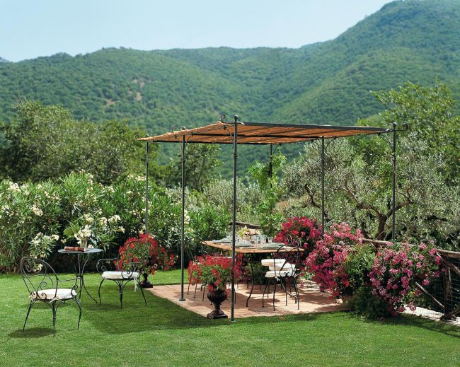Eine ruhige Außenterrasse in einem üppigen Garten mit einer Berglandschaft im Hintergrund. Die Terrasse verfügt über eine freistehende lineare Pergola Solaire mit Holzdach, einen runden Esstisch mit Stühlen und leuchtend blühende Pflanzen, die den Bereich umgeben. Zusätzliche Sitzgelegenheiten von Solaire befinden sich auf dem Rasen.