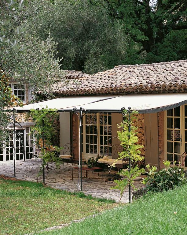 Ein charmantes Steinhaus mit Ziegeldach, eingebettet in üppiges Grün. Das Haus verfügt über große Glastüren, die auf eine gemütliche Terrasse mit schmiedeeisernen Stühlen und einem Tisch führen. Die Terrasse verfügt über eine gewölbte Anbaupergola Solaire, die Schatten spendet, und der umliegende Garten ist wunderschön angelegt.