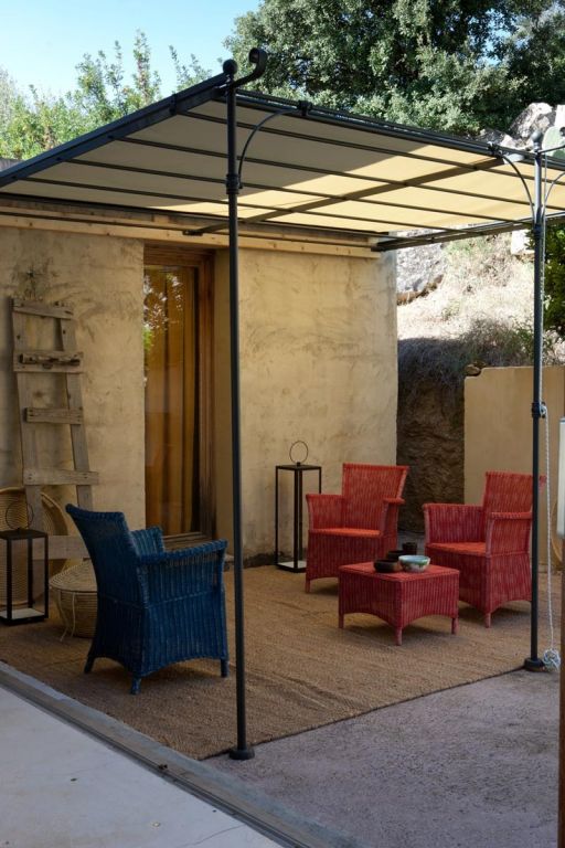 Sitzbereich im Freien mit einer überdachten Lineare Anbaupergola Solaire. Der Raum verfügt über einen Korbstuhl, eine Bank und einen kleinen Holztisch. Vier bunte Korbstühle (zwei blaue und zwei rote) stehen um einen kleinen runden Tisch auf einem gewebten Teppich. Eine Holzleiter lehnt an der Wand und trägt zum rustikalen Charme bei.