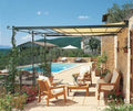 Eine malerische Außenterrasse mit Holzmöbeln und weißen Kissen wird von einer linearen Anbaupergola Solaire beschattet. Von der Terrasse aus blickt man auf einen Swimmingpool mit Liegestühlen und Sonnenschirmen. Um das Gebiet herum befinden sich Steingebäude, grüne Hügel und leuchtend blühende Pflanzen.