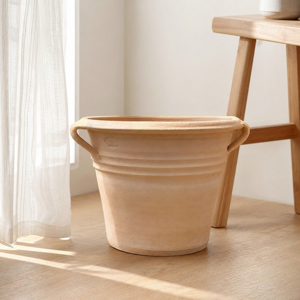 Der Terrakotta-Pflanzkübel „Sorbus“ mit Henkeln (Durchmesser 40–60 cm) in hellbrauner Ausführung steht auf einem Holzboden in der Nähe eines Fensters mit weißen, durchsichtigen Vorhängen und neben einem Holzhocker in einem sonnendurchfluteten Raum.