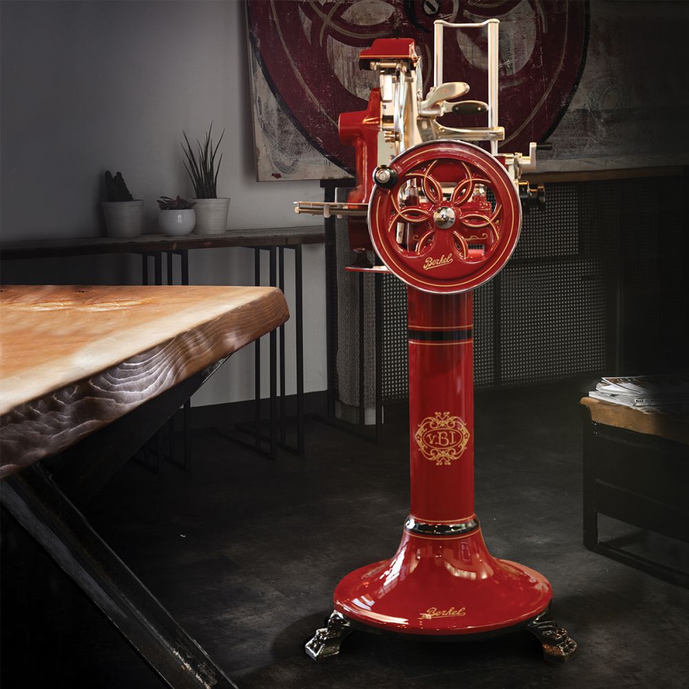 Eine markante Berkel Volano B2 Schwungradmaschine in Rot steht prominent in einem schwach beleuchteten Raum mit industriellem Dekor. Dieser Fleischschneider im Vintage-Stil verfügt über exquisite Messingdetails und ein dekoratives Emblem auf der Basis. Links ist ein einzigartiger Holztisch zu sehen, begleitet von Regalen, die mit Topfpflanzen und verschiedenen Küchenutensilien im Hintergrund geschmückt sind.