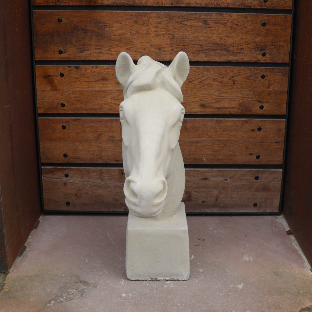 Die handgefertigte Steinfigur "Pferd groß" steht auf einem quadratischen Sockel vor einer Holzvertäfelung mit Metallbolzen, der Boden ist aus Beton und die Szenerie erinnert an einen Außenbereich.
