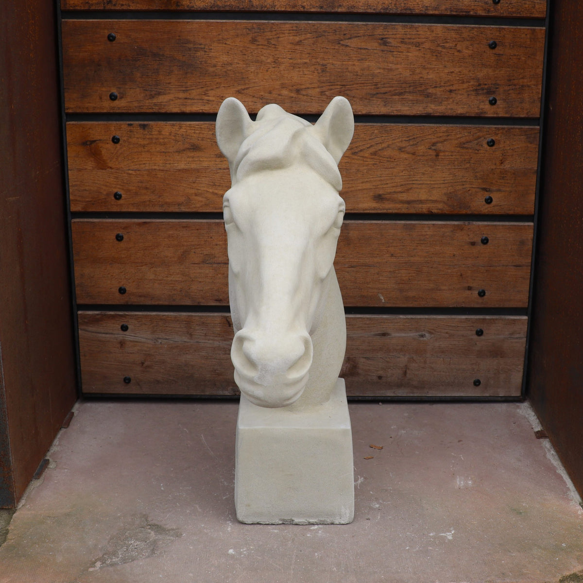 Die handgefertigte Steinfigur "Pferd groß" steht auf einem quadratischen Sockel vor einer Holzvertäfelung mit Metallbolzen, der Boden ist aus Beton und die Szenerie erinnert an einen Außenbereich.