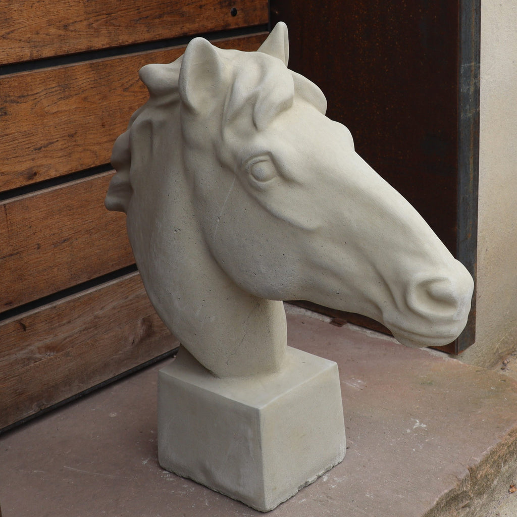 Die handgefertigte Steinfigur "Pferd groß" steht im Freien auf einer rechteckigen Steinsockel-Fläche nahe Holz- und Metallwänden.