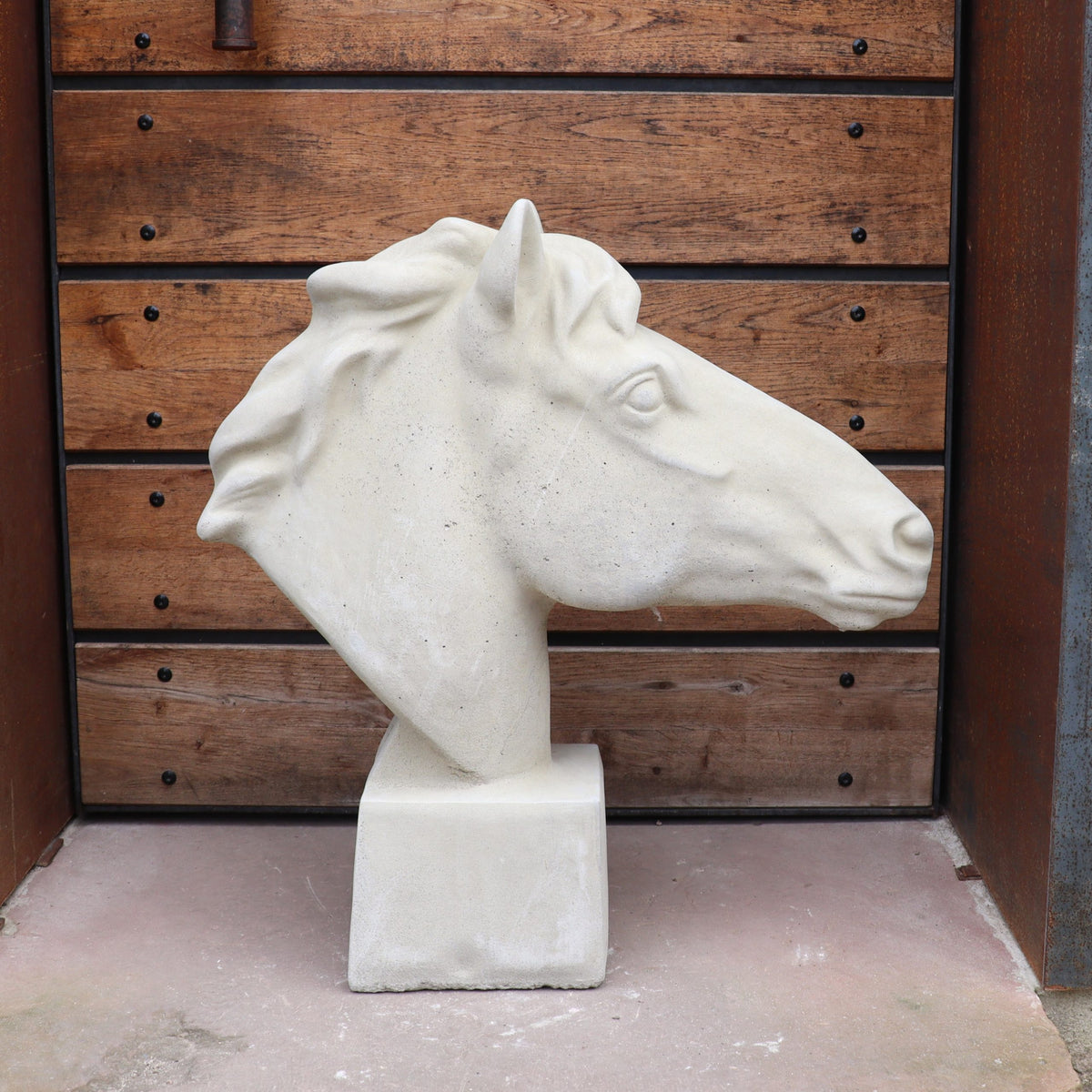 Die handgefertigte Steinfigur "Pferd groß" aus weißem Stein steht auf quadratischem Sockel vor einer Holztür mit Metallelementen.