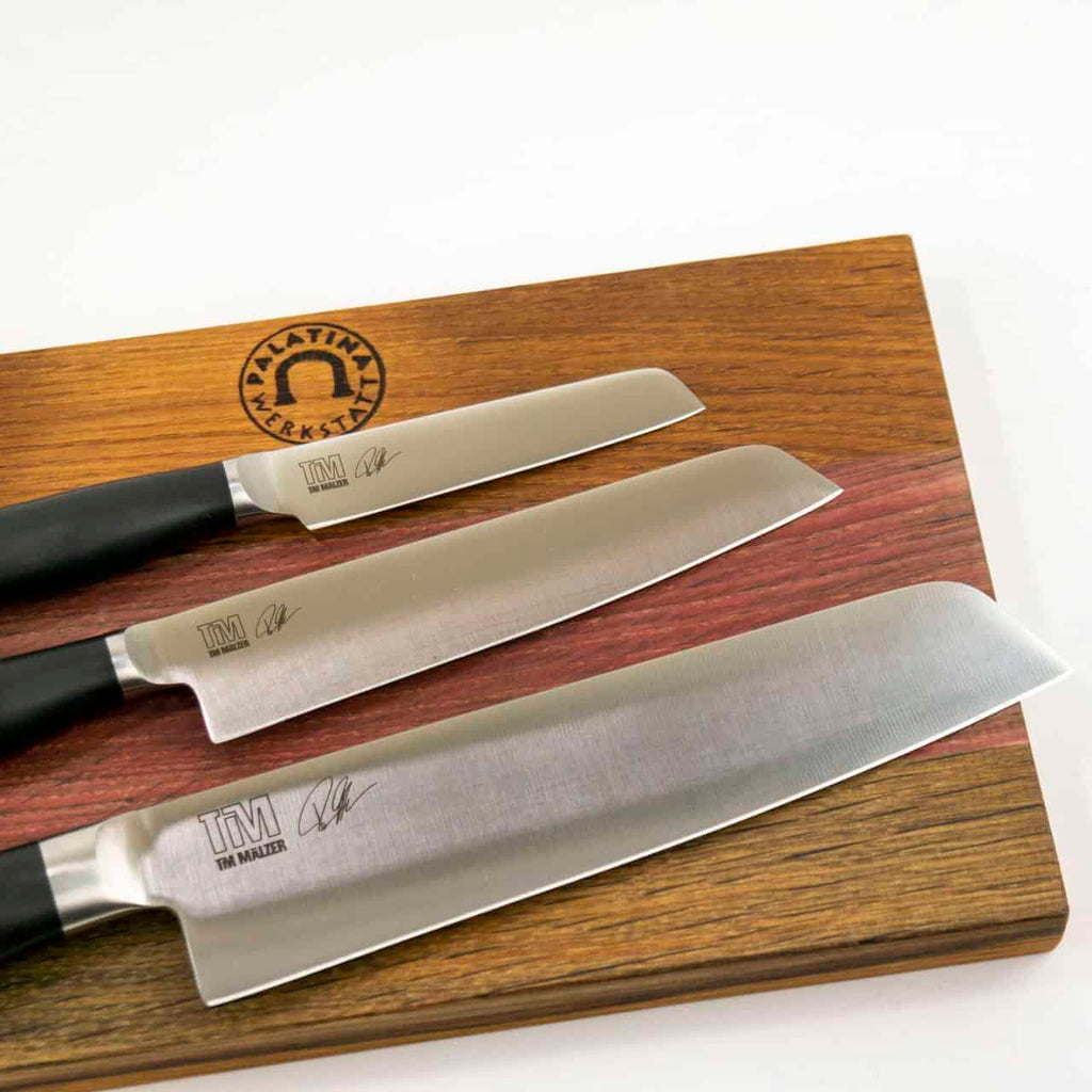 Ein Kai Shun Tim Mälzer Kamagata Geschenkset, bestehend aus drei Küchenmessern mit schwarzen Griffen auf einem massiven 30x18 cm großen Holzschneidebrett. Die Messer tragen Tim Mälzers Unterschrift und das „TM“-Logo, das die Marke repräsentiert. Das Schneidebrett ist mit einem Emblem mit dem Text „PALATINA LERREN“ und einem Hufeisensymbol verziert, wodurch ein exquisites Geschenkset entsteht.
