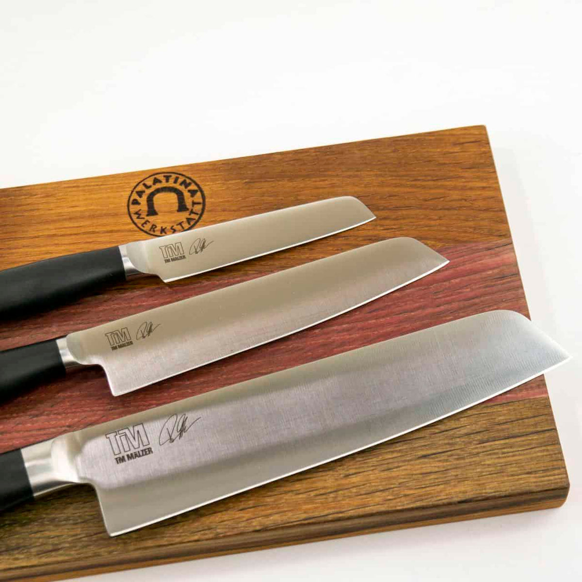 Ein Kai Shun Tim Mälzer Kamagata Geschenkset, bestehend aus drei Küchenmessern mit schwarzen Griffen auf einem massiven 30x18 cm großen Holzschneidebrett. Die Messer tragen Tim Mälzers Unterschrift und das „TM“-Logo, das die Marke repräsentiert. Das Schneidebrett ist mit einem Emblem mit dem Text „PALATINA LERREN“ und einem Hufeisensymbol verziert, wodurch ein exquisites Geschenkset entsteht.