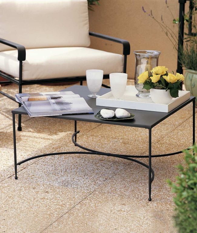 Eine gemütliche Szene auf einer Außenterrasse zeigt den Couchtisch quadratisch Toscana, einen Couchtisch aus Metall, auf dem ein offenes Buch, ein weißes Tablett mit einer Glaskanne, zwei Gläser und eine Vase mit gelben Rosen stehen. Daneben steht ein gepolsterter Sessel. Zwei Steine ruhen auf einer kleinen Schale in der Mitte des Couchtischs quadratisch Toscana und verstärken das toskanische Ambiente.