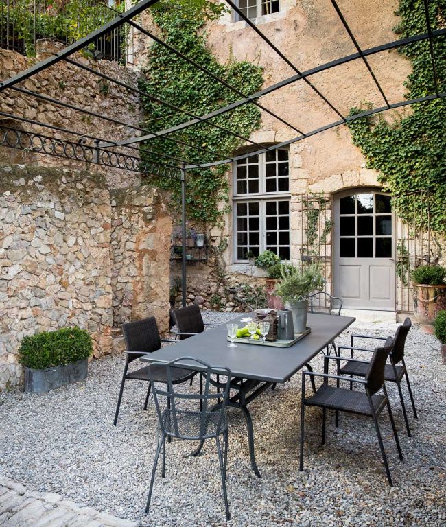 Eine rustikale Außenterrasse mit einer Pergola mit Metallrahmen verfügt über den rechteckigen Tisch Toscana (220 cm), der mit Topfpflanzen und einem Tablett geschmückt ist und von schwarzen Stühlen umgeben ist. Der Hintergrund besteht aus einer teilweise mit Efeu bewachsenen Steinmauer und einer Tür mit Glaseinsätzen, die den Charme der Toskana heraufbeschwört.