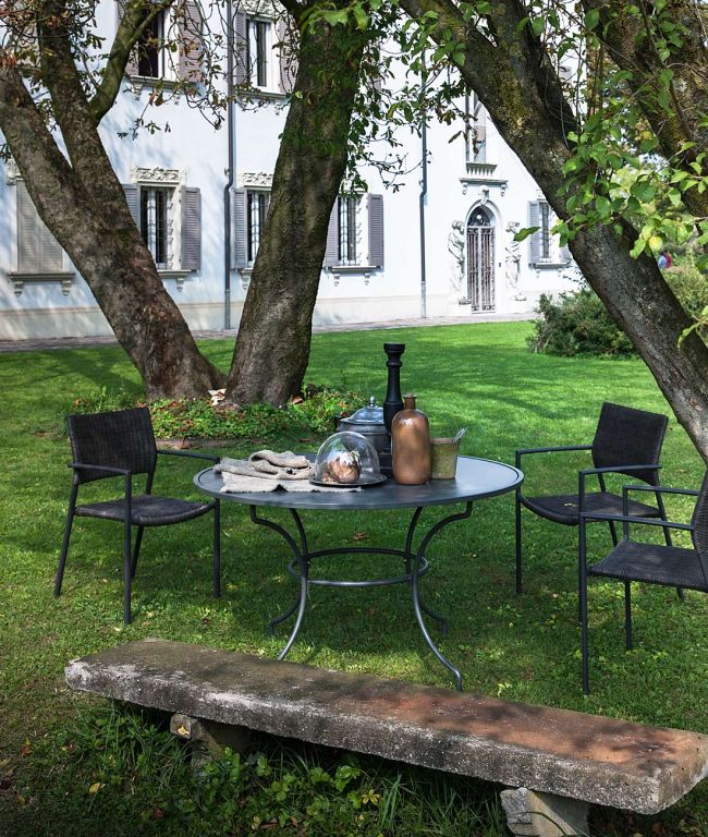 Eine ruhige Gartenszene mit einem Tisch Toscana rund 125 cm und zwei Stühlen unter großen Bäumen. Auf dem runden Tisch stehen dekorative Vasen, eine Glasglocke und ein Keramikkrug. Im Vordergrund steht eine Steinbank und im Hintergrund ein elegantes Gebäude mit Bogenfenstern, das an die Toskana erinnert.