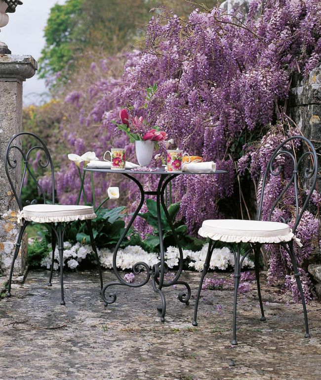 Ein charmanter Outdoor-Metalltisch und zwei Stühle mit weißen Kissen sind vor einem Hintergrund aus üppig blühenden lila Glyzinien angeordnet. Der runde Tisch rund Aurora Ø cm 70 ist mit einer Vase mit leuchtenden Blumen, zwei Teetassen und Tellern dekoriert und ruft eine gemütliche und einladende Gartentee-Darbietung hervor, die an einen Morgen im Licht der Aurora erinnert.