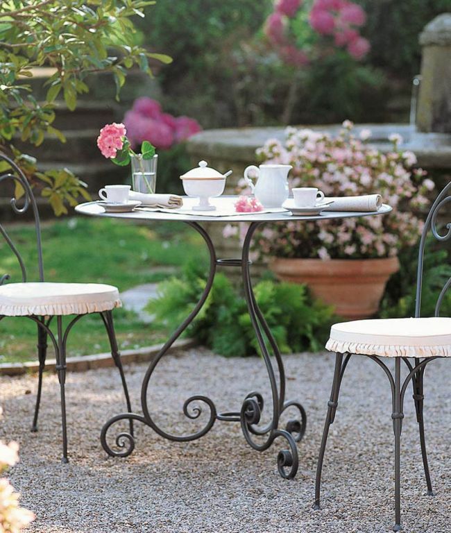Der Tisch rund Aurora Ø cm 90, ein runder Metalltisch mit eleganten, geschwungenen Beinen, ist mit einer Teekanne, Tassen und einer Vase mit rosa Blumen wunderschön für den Tee gedeckt. Passende gepolsterte Stühle umgeben den Tisch und vervollständigen die Szene auf einer Kiesterrasse, die im sanften Schein der Morgenröte mit blühenden rosa und grünen Pflanzen geschmückt ist.