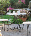 Der Tisch rund Aurora Ø cm 90, ein runder Metalltisch mit eleganten, geschwungenen Beinen, ist mit einer Teekanne, Tassen und einer Vase mit rosa Blumen wunderschön für den Tee gedeckt. Passende gepolsterte Stühle umgeben den Tisch und vervollständigen die Szene auf einer Kiesterrasse, die im sanften Schein der Morgenröte mit blühenden rosa und grünen Pflanzen geschmückt ist.