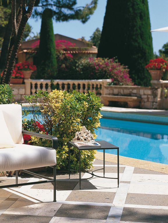 Eine ruhige Szene im Freien mit einer Terrasse mit Steinfliesen neben einem glitzernd blauen Pool. Ein weißer gepolsterter Stuhl und ein rechteckiger Couchtisch Toscana (52 x 38) mit Dekorationsgegenständen stehen neben leuchtend grünen Büschen und bunten Blumentöpfen und rufen den Charme der Toskana hervor. Im Hintergrund sind hohe Bäume und Sträucher zu sehen.