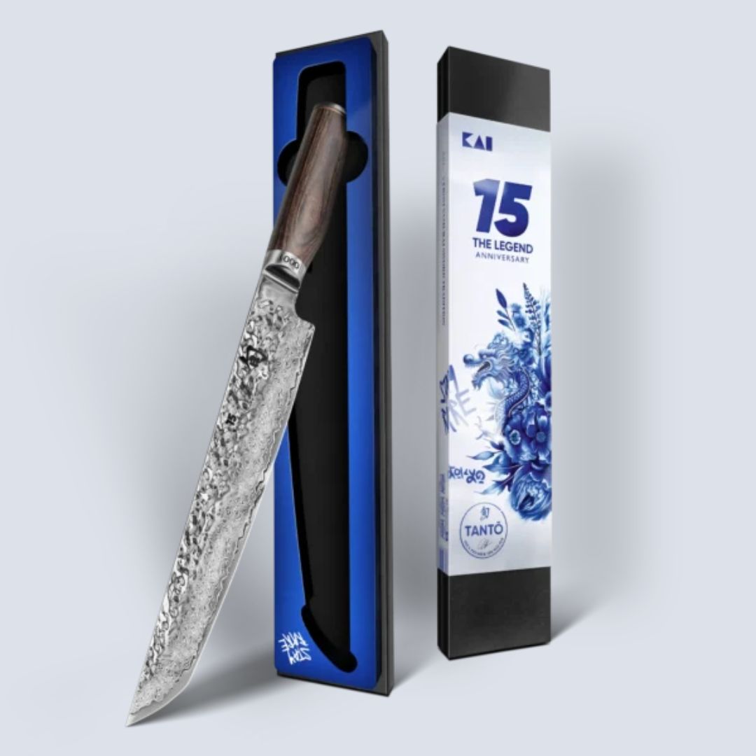 Ein Kai Shun Jubiläumsmesser TDM-1785 Tantō - 15 Jahre Tim Mälzer | 6.666 Stück aus gehämmertem Stahl mit dunklem Holzgriff wird in einer offenen schwarz-blauen Schachtel neben einer weißen Jubiläumsverpackung mit blauem Blumendruck und dem Text "15 The Legend" präsentiert.