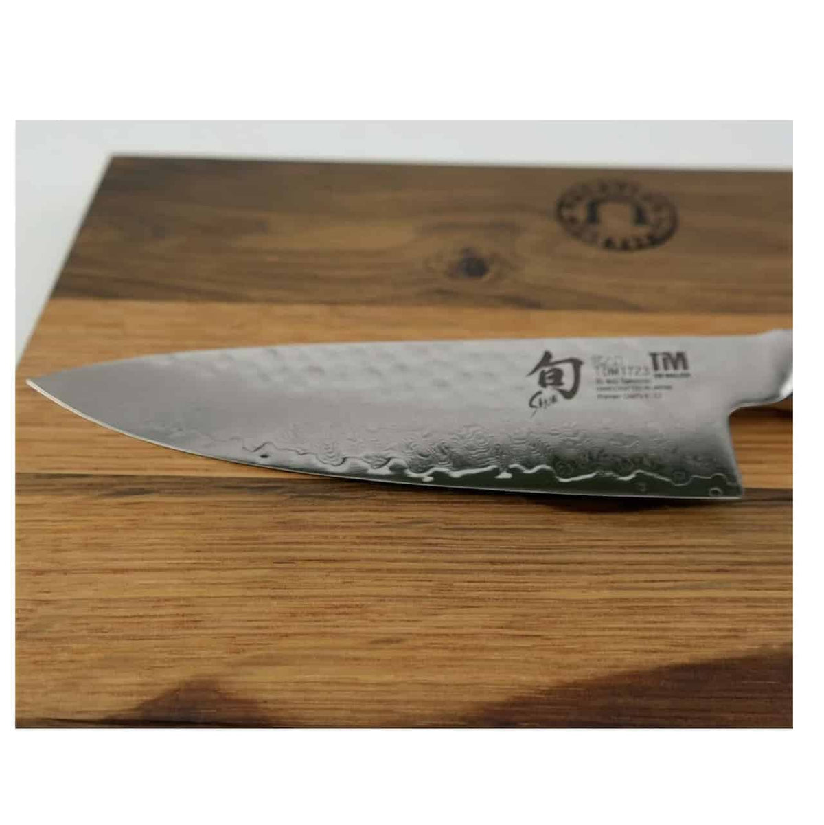 Eine Nahaufnahme eines glänzenden 15 cm langen Kai Shun Premier Tim Mälzer TDM-1723 Kochmessers mit gehämmerter Stahlstruktur, das auf einem hölzernen Küchenbrett Fassholz 30 x 18 cm mit dunkler Holzmaserung ruht. Das Messer weist einige Inschriften in der Nähe des Klingengriffs auf, die seine Marke und sein Modell angeben.