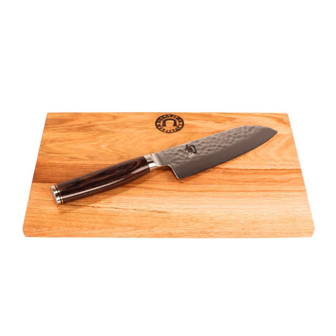 Das Kai Shun Premier Tim Mälzer kleines Santoku (14 cm) mit dunklem Holzgriff und gehämmerter Klinge ruht auf dem mitgelieferten 30x20 cm großen Schneidebrett mit kreisförmigem Logo am oberen Rand.