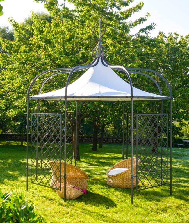 Ein achteckiger Pavillon Solaire mit raffiniertem Metallrahmen und elegantem Spitzdach steht auf einem üppigen grünen Rasen. Im Inneren sind zwei geflochtene Rattanstühle mit rosa Kissen nebeneinander angeordnet und einander zugewandt. Umgeben von Bäumen schafft dieser achteckige Pavillon einen ruhigen und schattigen Ort.
