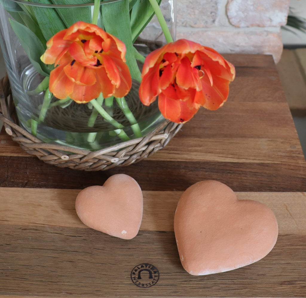 Eine Vase mit leuchtend orangefarbenen Tulpen steht auf einem Holztisch neben zwei handgefertigten Deko-Herzen aus Terrakotta, frostfesten Tonornamenten. Der Tisch hat einen rustikalen Charme mit einem gewebten Tischset unter der Vase. Im Hintergrund ist eine Backsteinmauer, die der Szene eine gemütliche und doch festliche Frostfest-Atmosphäre verleiht.