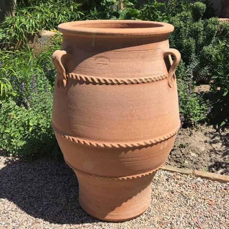 Die frostfeste Terrakotta-Amphore „Thymus“ ist 70 cm hoch und mit dekorativen Bändern sowie zwei Henkeln an jeder Seite versehen. Sie steht auf einer Kiesfläche, umgeben von Grünpflanzen, die an eine Gartenanlage im Freien erinnern.
