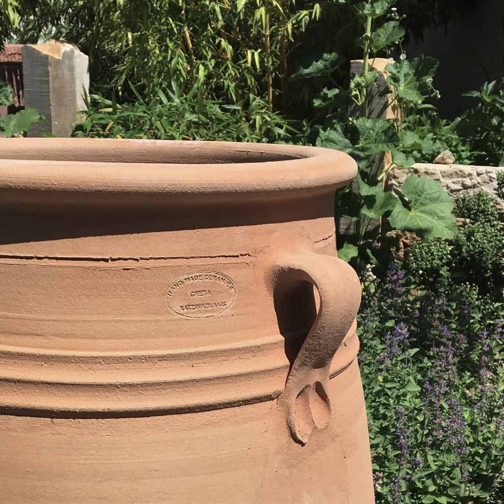 Das Amphore Garten Winterfest "Thymus" H=70cm, mit Henkel und Stempel "HAND MADE GREECE", steht im Freien zwischen grünen Pflanzen und lila Blumen und verleiht Ihrem Garten das ganze Jahr über Charme.