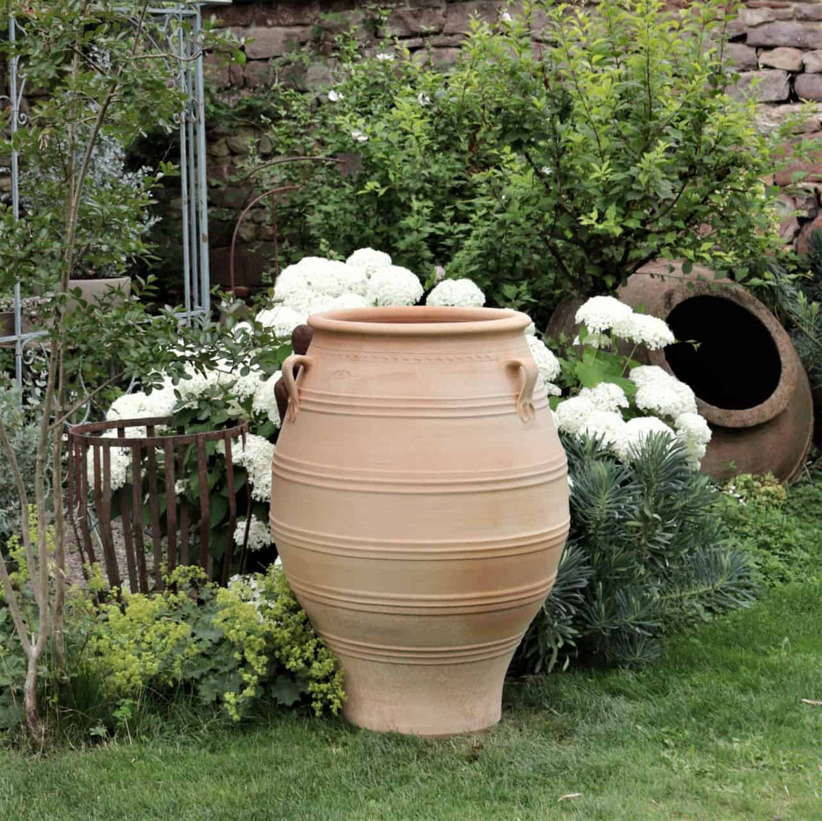 Ein großer, beiger Keramiktopf steht in einem üppigen Garten, umgeben von grünem Laub und weiß blühenden Pflanzen. Ein alter, kleinerer Topf liegt auf der Seite im Hintergrund neben einer Steinmauer und trägt zum rustikalen Charme der Umgebung bei, während eine XXL-Terrakotta-Amphore „Thymus“ mit einer Höhe von 100 cm inmitten des Grüns steht.