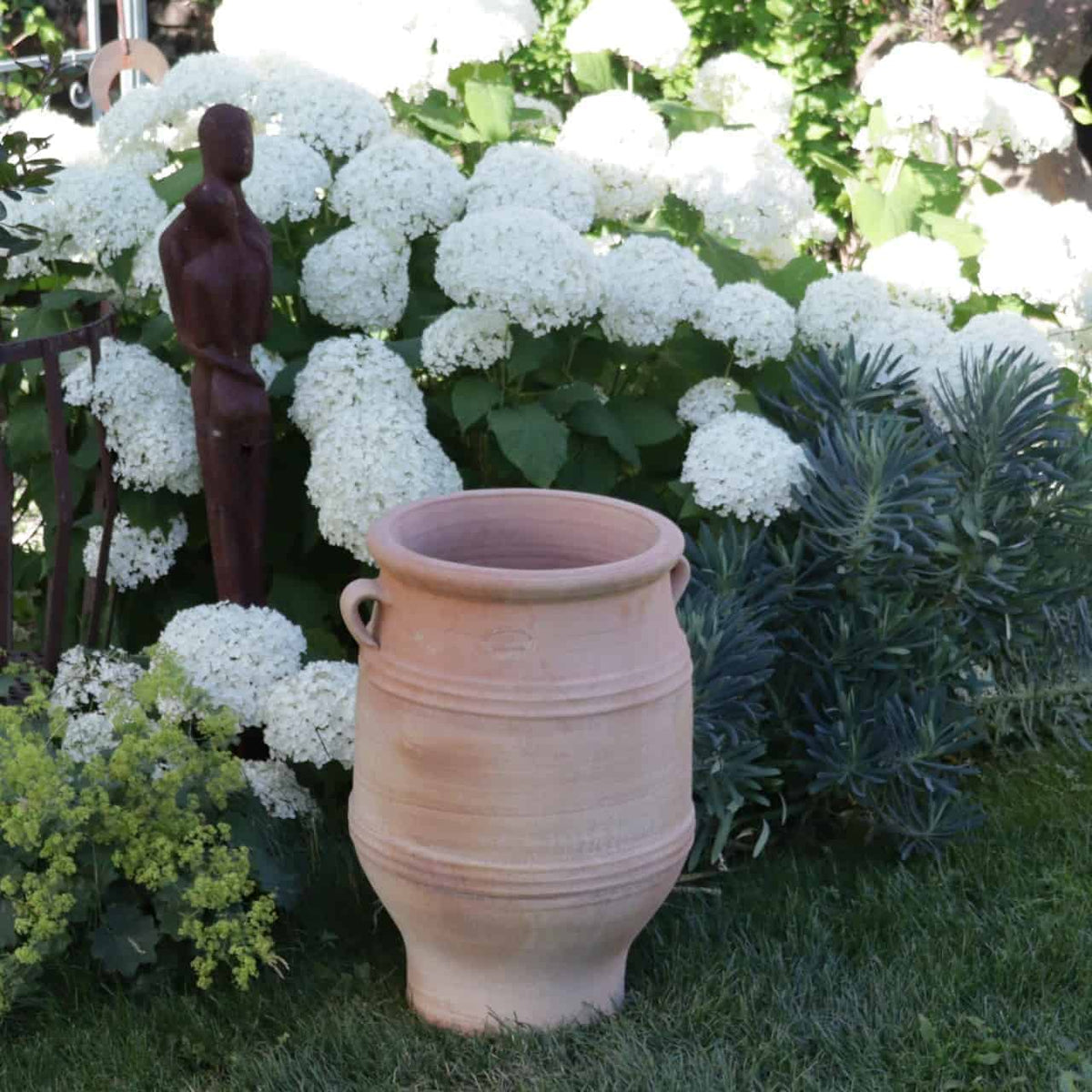 Ein frostfester Amphore Garten „Thymus“, 50 cm hoch, steht auf dem Gras vor einem üppigen Garten mit weißen Hortensien in voller Blüte. Neben dem Topf steht eine hohe, schlanke, abstrakte Bronzeskulptur einer menschlichen Figur. Grünes Laub und blühender Thymus umgeben das Exponat und schaffen eine ruhige Gartenszene.