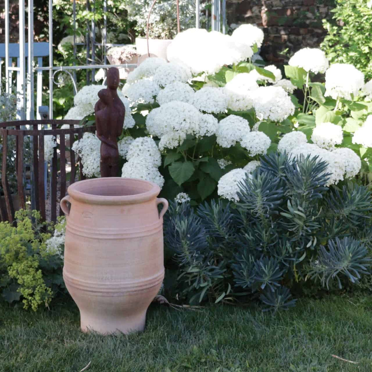 Eine Gartenszene mit großen weißen Hortensienblüten in voller Blüte. Davor steht ein hoher, frostfester Amphore Garten „Thymus“, H=50cm, flankiert von einer Holzskulptur und üppigen grünen Pflanzen. Im Hintergrund sind ein schmiedeeiserner Zaun und ein Zaunpfahl zu sehen, die mit durchscheinenden Thymusblüten einen rustikalen Charme verleihen.