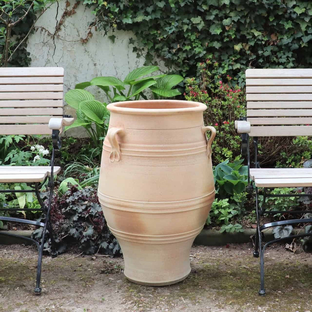 Das beige Keramik-Amphore Garten Winterfest „Thymus“ H=70cm mit zwei Griffen steht zwischen hölzernen Gartenbänken, umgeben von üppigen Pflanzen und Efeu und schafft eine heitere Außenatmosphäre.